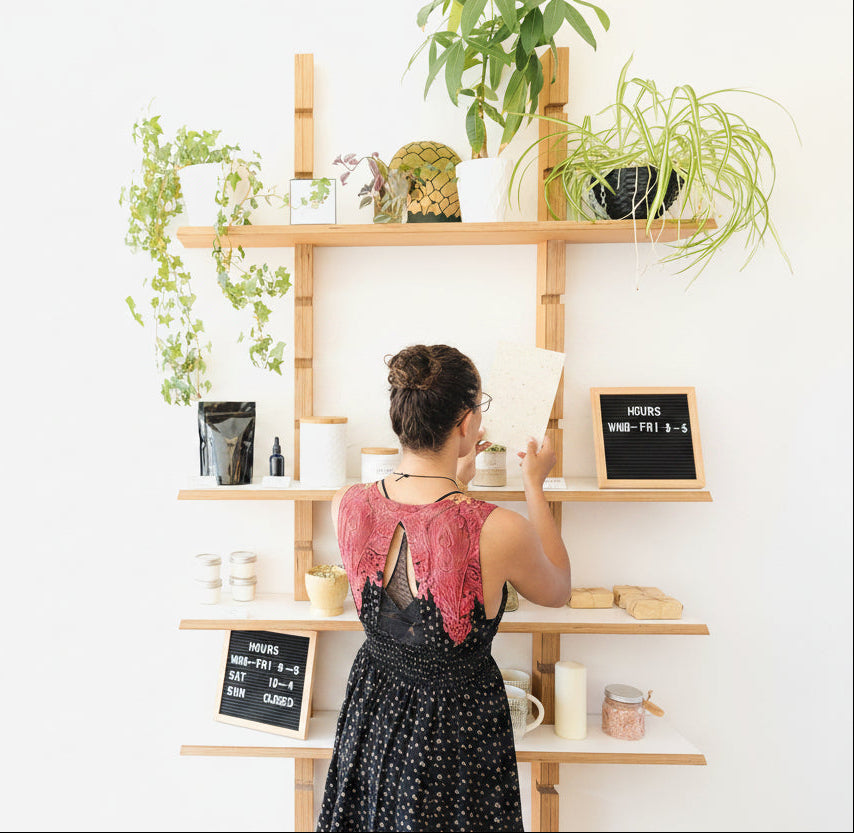 A woman in a black and pink dress stands with her back to the camera, holding a seed paper sheet in front of a tall wooden shelving unit decorated with plants and small decor items against a white wall.
